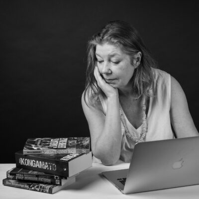 Mikala Rosenkilde ser ud til at være fordybet i tanker, mens hun sidder ved et skrivebord med en åben bærbar computer foran sig og en stak bøger ved hendes side. Billedet er i sort-hvid.